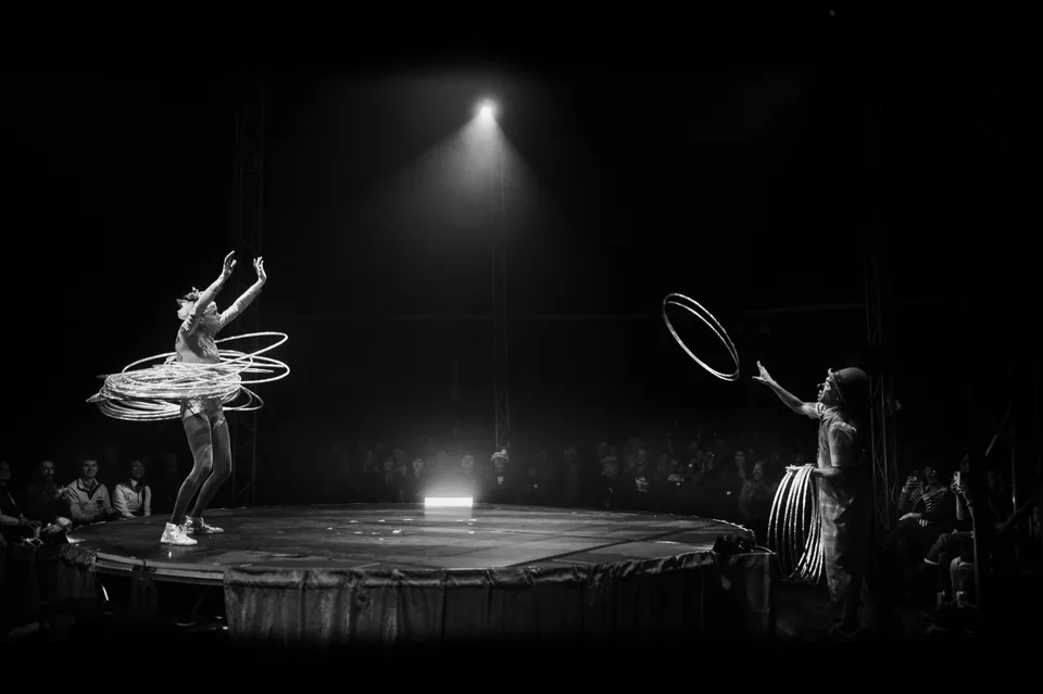 Two circus performers exchange hula hoops across a dark stage.