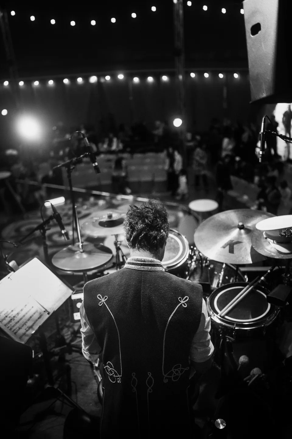 A drummer sits at a drum kit facing stage lights and an audience.